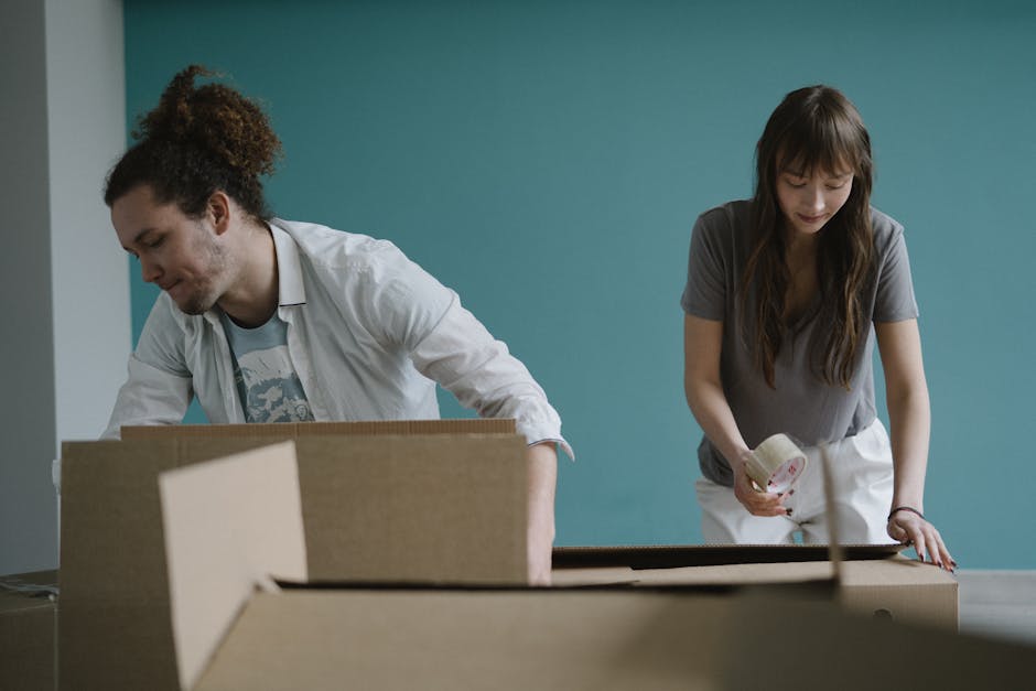 A man in a grey hoodie is bending over to pick up or place a cardboard box labeled 'pillows' inside a room with natural light coming through a large window with grey curtains. Several other cardboard